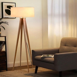 May include: A wooden tripod floor lamp with a white fabric lampshade. The lamp is in a living room setting with a chair and a bookshelf.