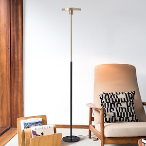 May include: A modern floor lamp with a black and gold finish. The lamp has a circular base, a black pole, and a gold-colored upper section with a flat, circular light. The lamp is next to a wooden chair and a magazine rack.