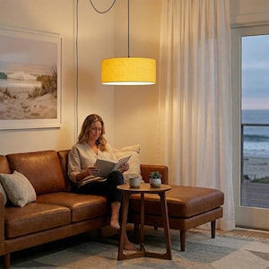May include: A yellow cylindrical pendant lamp hangs above a brown leather sofa. The lamp is suspended from a black cord. A woman is sitting on the sofa reading a book. A small wooden side table and a window with a view of the ocean are also visible.