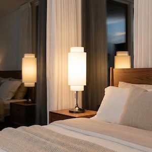 May include: A modern bedroom scene featuring a bed with white bedding and a beige throw blanket. Two bedside tables each hold a cylindrical lamp with a white shade. The lamps have a black base and emit a warm glow, enhancing the room's ambiance.