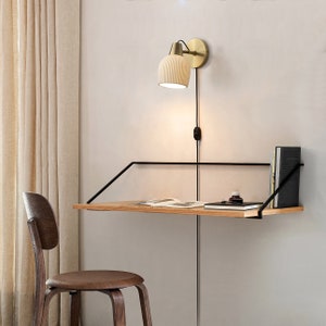 May include: A wall-mounted desk with a wooden surface and black metal supports. A brass-colored lamp is mounted above the desk. A wooden chair is in front of the desk. Books and a small bottle are on the desk.