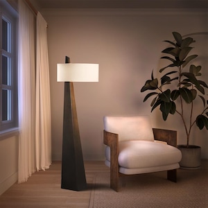 May include: A modern floor lamp with a geometric, dark bronze base and a rectangular off-white lampshade. The lamp stands tall next to a cream-colored armchair with wooden arms and a potted plant, creating a cozy interior design.