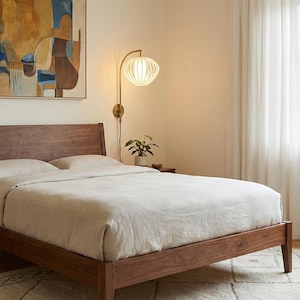 May include: A bedroom scene featuring a wooden bed frame with a light-colored duvet and pillows. A wall-mounted lamp with a white, ribbed shade and a brass arm is positioned above the bed. A small potted plant sits on a nightstand.