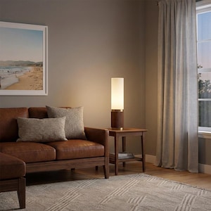 May include: A modern table lamp with a cylindrical white shade and a dark wood base with brass accents. The lamp sits on a small wooden side table next to a brown leather sofa. A framed beach scene hangs on the wall.