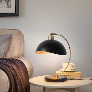 May include: A black desk lamp with a curved, brass-colored arm and a rounded shade. The interior of the shade is gold. The lamp sits on a round base and is next to a stack of books and a cup of coffee on a small, white table.