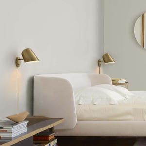 May include: A bedroom scene featuring a bed with a light-colored headboard and white bedding. Two brass wall-mounted lamps are positioned above the bed. A small side table holds books and a decorative object.