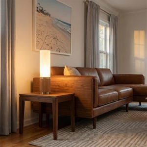 May include: A modern living room scene featuring a wooden side table with a cylindrical lamp. The lamp has a white shade and a wooden base. A brown leather sofa and a framed beach scene complete the room's decor.