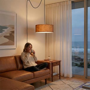 May include: A brown leather sofa with a woman sitting on it, holding a mug. A pendant light with a brown shade hangs above. A small wooden side table holds a small white vase and a coaster. The room has a view of the ocean.