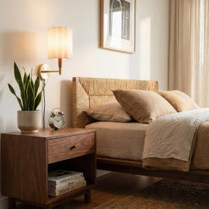 May include: A bedroom scene featuring a wooden nightstand with a drawer and open shelf, holding books and a potted plant. A bed with a woven headboard and neutral-toned bedding is in the background. A wall-mounted lamp and an alarm clock are on the nightstand.