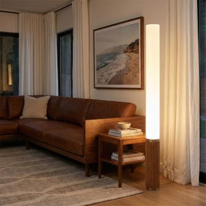 May include: A modern living room featuring a brown leather sectional sofa, a wooden side table, and a tall, cylindrical floor lamp with a white shade. A framed landscape art piece hangs on the wall, and white curtains frame the window.