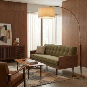May include: A tall, arched floor lamp with a brass-finished metal stand, a wooden accent section, and a large, cylindrical, pleated off-white fabric shade that provides overhead lighting for a room with a green sofa and wooden furniture.