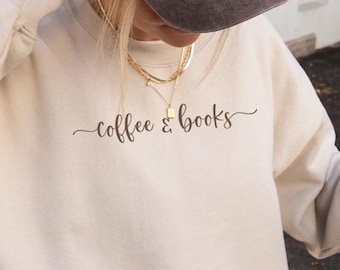 May include: A woman wearing a cream-colored sweatshirt with the text "coffee & books" printed on the front. She is holding a clear plastic cup with a brown beverage.