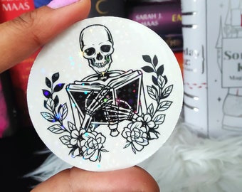 May include: A white sticker with a black and white illustration of a skeleton reading a book. The book is open and the skeleton is holding it with both hands. The sticker has a glittery finish and is surrounded by black floral designs.