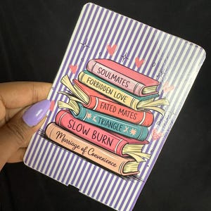 May include: A rectangular card with a stack of books illustration. The books are in various colors and have titles like "Soulmates" and "Forbidden Love." The background has vertical purple and white stripes, with small hearts and stars.