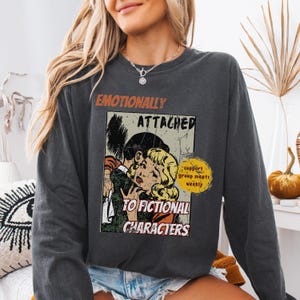 May include: Dark gray sweatshirt with the text "Emotionally Attached to Fictional Characters" and a vintage comic book-style graphic of a couple embracing. The sweatshirt also has a yellow circle with the text "support group meets weekly".