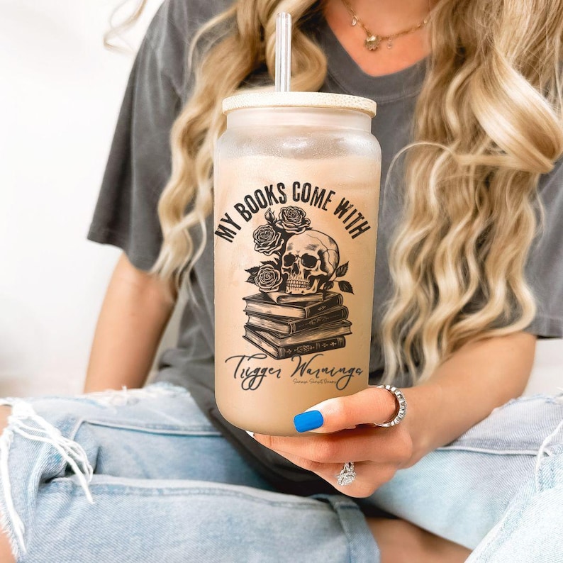 may include; a frosted glass with a bamboo lid that has a black graphic of a stack of books with a skull resting on top. There are also roses included in the design by the skull. The text on the design reads 'my books come with trigger warnings'