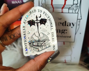 May include: A white sticker with a black dragon and a stack of books. The sticker has a holographic finish and the text "Emotionally Attached to Fictional Dragons".