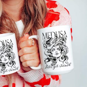 May include: A white ceramic mug featuring a black graphic illustration of Medusa with snakes for hair. The text MEDUSA is printed above the illustration, and the phrase Lets get stoned is printed in a cursive script font below the image.