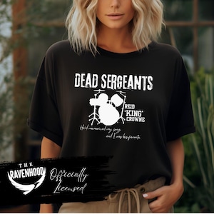 Peut inclure: Un t-shirt noir à manches courtes avec le texte blanc DEAD SERGEANTS, REID KING CROWNE, et He'd memorized my song, and I was his favorite, ainsi qu'un graphique blanc d'une batterie et le logo THE RAVENHOOD Officially Licensed.