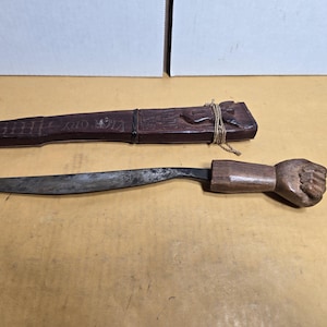May include: A vintage knife with a wooden handle carved in the shape of a fist, and a wooden sheath. The blade is curved and has a dark, aged appearance. The sheath is dark brown and has carved details.