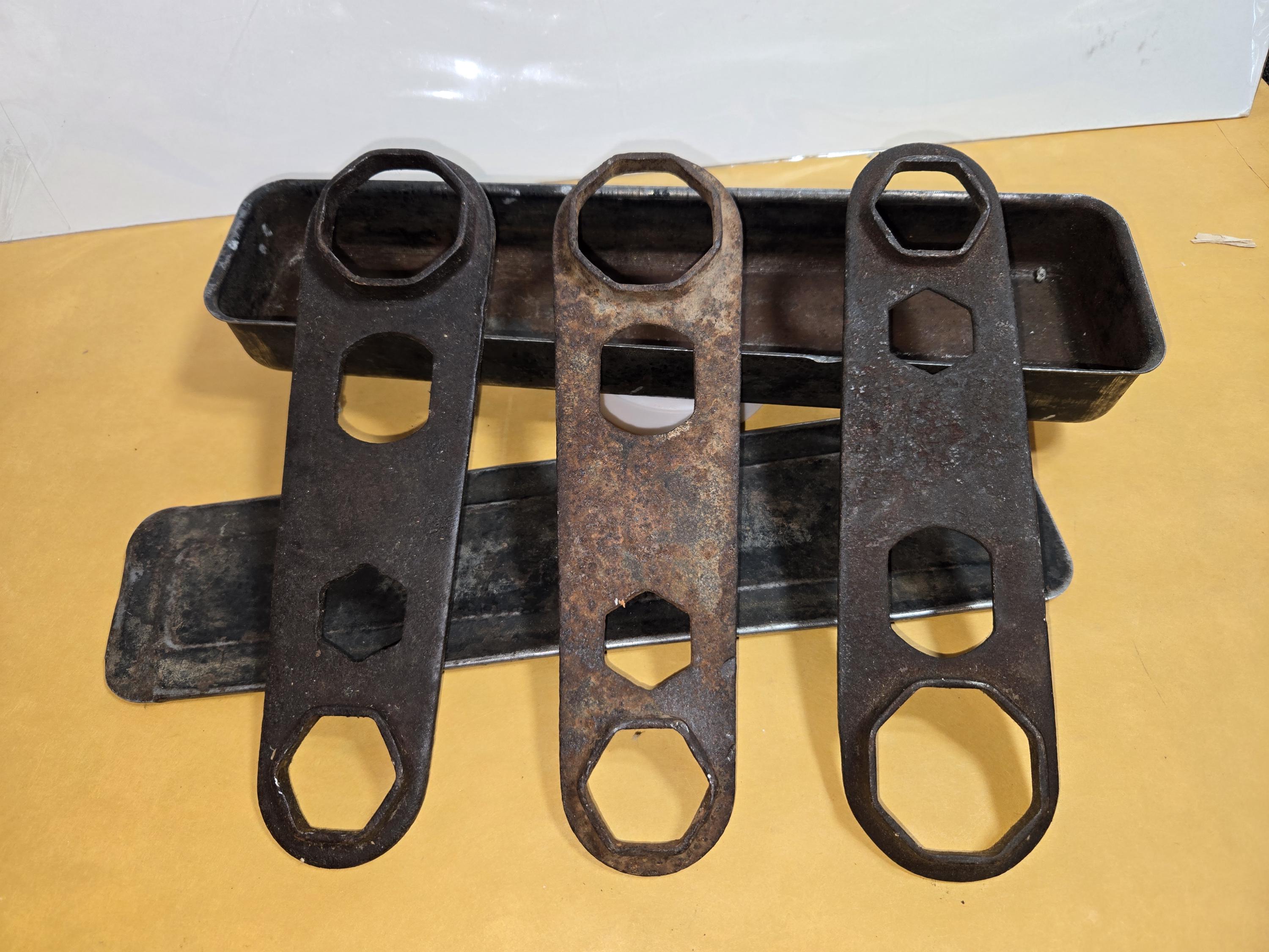 May include: Three dark, weathered metal multi-wrench tools with various hexagonal cutouts arranged vertically. Behind them sits a rectangular metal storage tray with a separate lid, showing signs of age, oxidation, and dark surface wear.