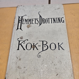 May include: A vintage cookbook titled "Hemmets Drottning Kok-Bok" in black script on a textured, off-white cover. The book shows signs of age with visible wear and discoloration. The cover has a dotted pattern.