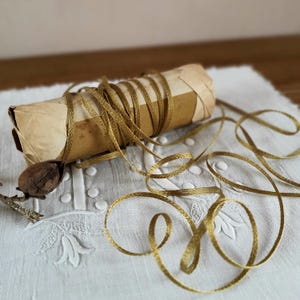 Può includere: Un rotolo di carta marrone avvolto attorno a un oggetto, con nastro dorato arrotolato attorno e che si estende verso destra. Il nastro è anche avvolto attorno al rotolo. Una piccola noce marrone e un rametto sono a sinistra.