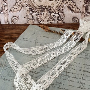 Peut inclure: Ruban de dentelle blanc cassé avec un motif floral délicat. Le ruban est disposé en lignes fluides sur un fond de papier vintage et un élément mural décoratif. La dentelle a un bord festonné.