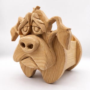 May include: A wooden dog sculpture with a mournful expression. The dog features large eyes, a prominent nose, and a small compartment. The sculpture is crafted from light-coloured wood, showcasing the natural grain.