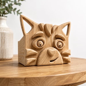 May include: A wooden cat-shaped box with a carved face, ears, and whiskers. The box is made of light-colored wood and sits on a round wooden table. The cat's face has large, round eyes and a smiling mouth. The box is a decorative item.