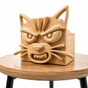 May include: A wooden box with a carved cat face design. The cat has an angry expression with furrowed brows, large eyes, and visible teeth. The box is made of light-colored wood and sits on a round wooden table with black legs.