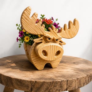 K&ouml;nnte beinhalten: Ein h&ouml;lzerner Elch-Blumentopf gef&uuml;llt mit bunten Blumen. Der Elch hat gro&szlig;e Geweihe und ein detailliertes Gesicht. Der Pflanztopf steht auf einem rustikalen Holzschemel und schafft eine charmante, nat&uuml;rliche &Auml;sthetik. Die Blumen umfassen gelbe, rote und violette Bl&uuml;ten.