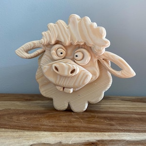 Może przedstawiać: Jasnobrązowa drewniana figurka owcy w stylu kreskówkowym. Owca ma duże oczy, szeroki pysk i puszyste, teksturowane futro. Uszy są zakrzywione. Drewniany przedmiot jest dekoracyjny i ma zabawny wygląd.