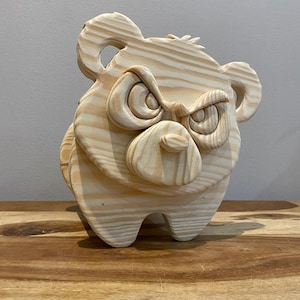 Puede incluir: Una escultura de madera de un oso con una expresión de enfado tallada. El oso está hecho de madera marrón claro y tiene un estilo simple y de dibujos animados.