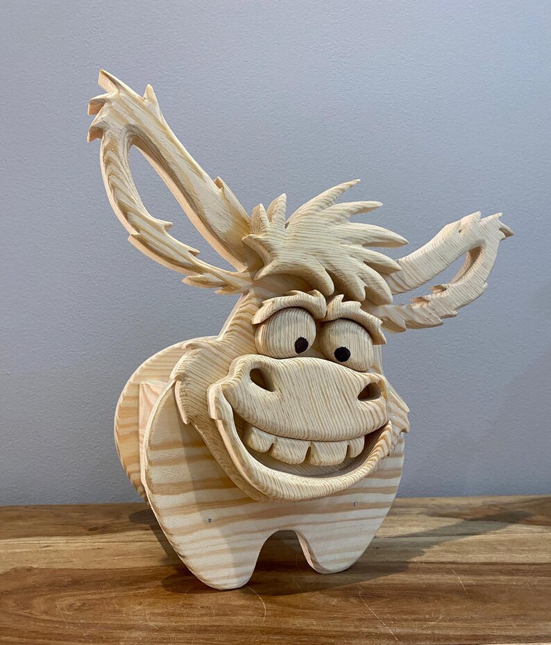 May include: A whimsical, wooden donkey sculpture with a large, smiling face and oversized ears. The donkey is carved from light-colored wood, showcasing the natural grain. The sculpture has a cartoonish design, perfect for home decor.
