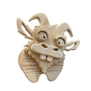 May include: A whimsical wooden sculpture of a cartoonish goat's head. The sculpture is made of light-colored wood, with visible grain. It features large, expressive eyes, a wide snout with teeth, and curved horns. The goat's head is mounted on a wooden base.