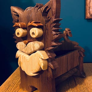 May include: A handcrafted wooden box in the shape of a cat. The box is made of dark brown wood with lighter wood accents for the eyes, nose, and beard. The cat's face is intricately carved, and the box has a storage compartment.