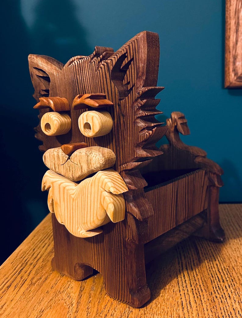 May include: A handcrafted wooden box in the shape of a cat. The box is made of dark brown wood with lighter wood accents for the eyes, nose, and beard. The cat's face is intricately carved, and the box has a storage compartment.