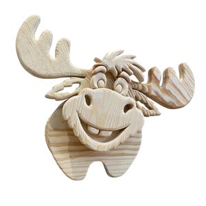 May include: A wooden moose head sculpture with a smiling face and large antlers. The sculpture is made of light-colored wood, with visible grain patterns. The moose has a cheerful expression, perfect for a rustic or woodland-themed decor.