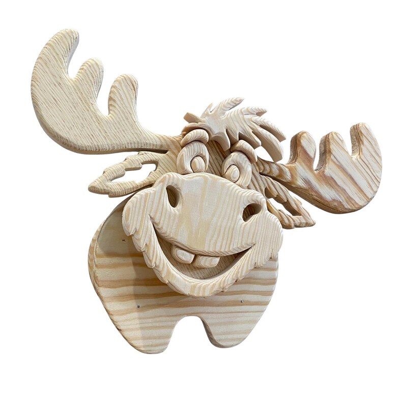 May include: A wooden moose head sculpture with a smiling face and large antlers. The sculpture is made of light-colored wood, with visible grain patterns. The moose has a cheerful expression, perfect for a rustic or woodland-themed decor.