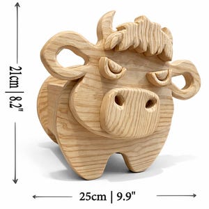 K&ouml;nnte beinhalten: Dekorative Kuh aus Holz. Die Kuh ist aus hellem Holz gefertigt und misst 21 cm in der H&ouml;he und 25 cm in der Breite. Sie weist geschnitzte Details wie H&ouml;rner, Ohren und ein stilisiertes Gesicht auf. Der Artikel ist zum Aufstellen konzipiert.
