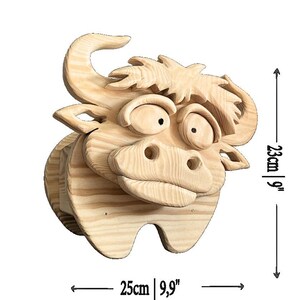 May include: A wooden bull head sculpture, measuring 9 inches high and 9.9 inches wide. The sculpture is made of light-colored wood and features a cartoonish design with large eyes and a prominent snout. The bull has a whimsical, friendly appearance.