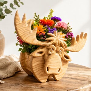 May include: A wooden moose-shaped planter filled with colorful flowers. The planter is made of light-colored wood and features a detailed carving of a moose's face and antlers. The flowers include orange, purple, pink, and yellow blooms, creating a vibrant display.