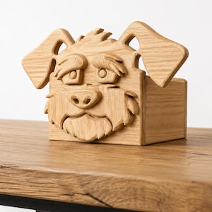 Puede incluir: Una caja de madera con un diseño tallado de cara de perro. La cara del perro presenta ojos, nariz y boca detallados, con grandes orejas que se extienden desde la parte superior. La caja es de madera clara y está sobre una superficie de madera.