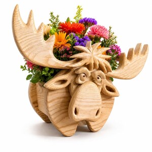 May include: A wooden moose planter with a carved face, antlers, and body. The planter is filled with a variety of colorful flowers, including orange, red, purple, and pink blooms. The moose is made of light-colored wood.