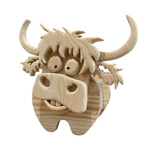May include: A whimsical, wooden Highland cow head sculpture. The sculpture is crafted from light-colored wood and features large, expressive eyes, a wide snout, and curved horns. The cow has a shaggy mane and a playful, cartoonish design.