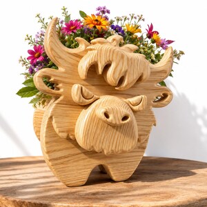 K&ouml;nnte beinhalten: Ein Holzblumentopf in Form einer Highland-Kuh, gef&uuml;llt mit bunten Blumen. Der Topf ist aus hellem Holz geschnitzt und weist detaillierte H&ouml;rner, eine Schnauze und eine zottelige M&auml;hne auf. Die Blumen umfassen rosa, gelbe und violette Bl&uuml;ten.