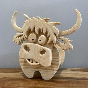 May include: A whimsical wooden sculpture of a Highland cow. The cow has large, expressive eyes, a wide grin, and curved horns. The sculpture is made of light-colored wood and has a textured, carved appearance. The cow is standing on a wooden surface.