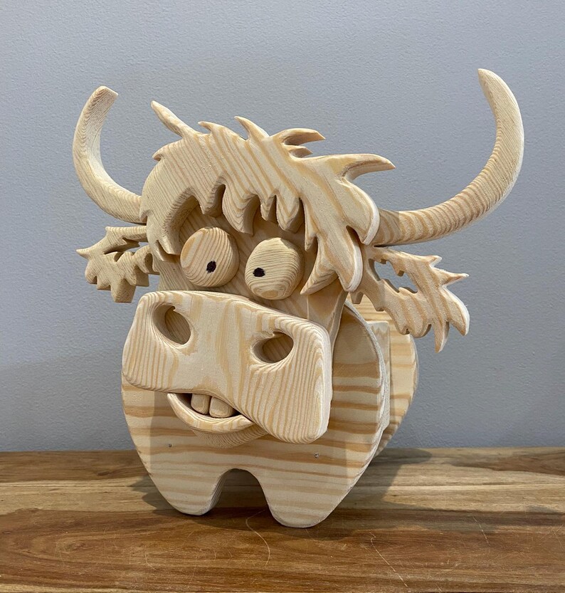 May include: A whimsical wooden sculpture of a Highland cow. The cow has large, expressive eyes, a wide grin, and curved horns. The sculpture is made of light-colored wood and has a textured, carved appearance. The cow is standing on a wooden surface.