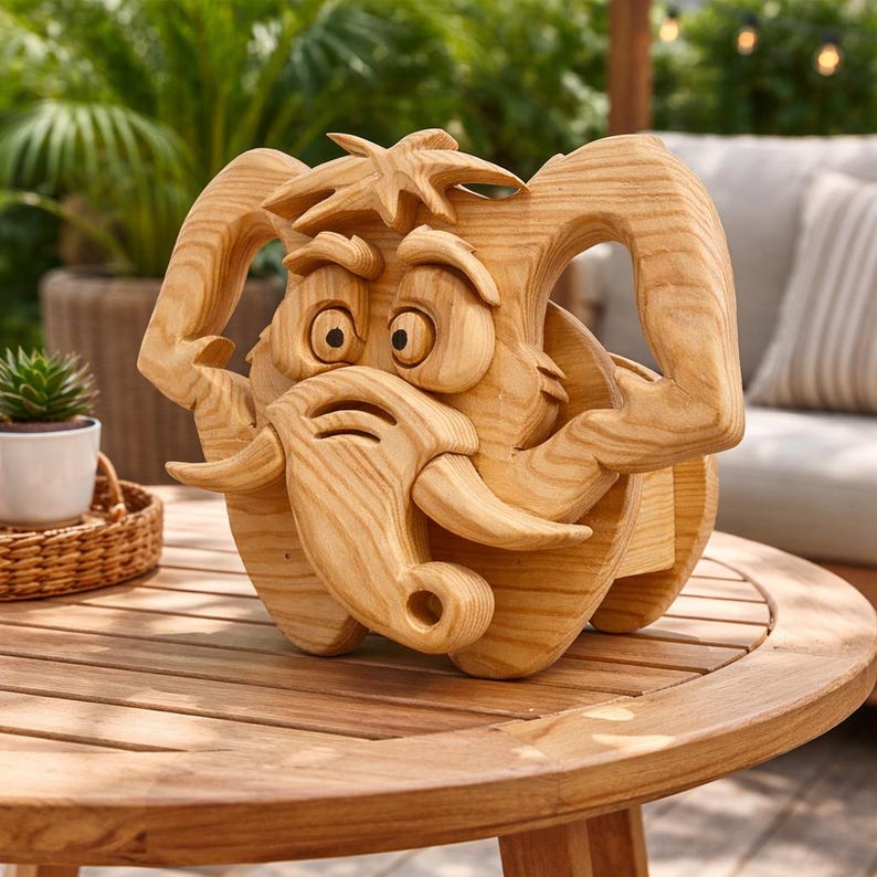 May include: A wooden sculpture of a cartoon elephant with large ears, tusks, and expressive eyes. The sculpture is made of light-colored wood and sits on a wooden table. The elephant has a whimsical design and is likely a decorative item.